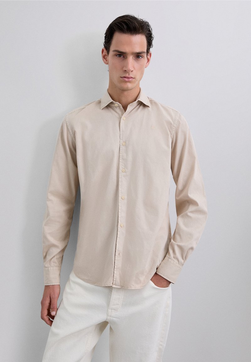 Camisa color beige claro de botones, hecha de algodón suave, con cuello, mangas largas y detalles de costura sutiles. Textura suave y corte regular.