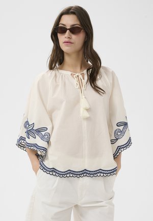 Vrouw draagt een loszittend wit blouse met marineblauwe borduur op de mouwen en zoom, gecombineerd met witte broek en smalle donkere zonnebril.