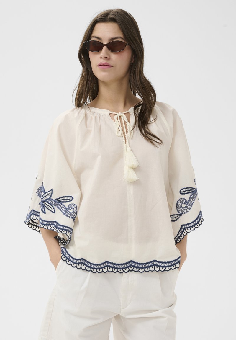 Femme portant un chemisier blanc ample avec une broderie marine sur les manches et l'ourlet, associé à un pantalon blanc et des lunettes de soleil étroites et foncées.
