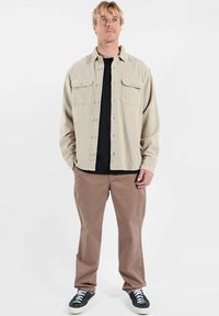 Chemise en velours côtelé beige avec deux poches poitrine, fermeture boutonnée à l'avant, portée par-dessus un t-shirt noir, associée à un pantalon marron clair et à des baskets noires.