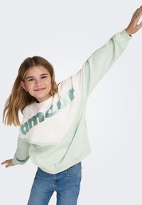 Μεντ πράσινο φούτερ με λευκή επιφάνεια, μεταλλικό πράσινο κείμενο «amour», μακριά μανίκια, ριμπ μανσέτες και άνετη εφαρμογή. Συνδυασμένο με μπλε τζιν.