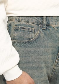 Light blue denim jeans with a faded wash, featuring a logo tag on the back pocket and a white textured sleeve visible at the side.