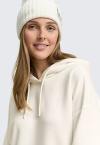 Mujer con un gorro blanco de canalé y una sudadera color crema con un pequeño logo en relieve, sonriendo con un maquillaje ligero y cabello largo rubio.