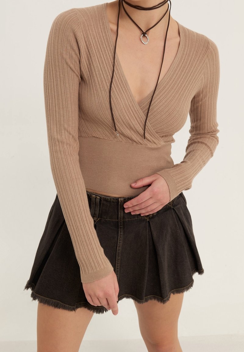 Femme portant un haut cache-cœur côtelé à manches longues beige et une mini-jupe en denim plissée noire, avec un collier en cordon noir orné d'un anneau en argent.