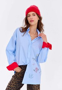 Kobieta w czerwonej czapce beanie, w za dużej jasnoniebieskiej koszuli z czerwonymi mankietami i szkicami, w zestawieniu z spodniami w panterkę, pozująca na tle białego tła.