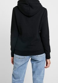 Felpa nera con cappuccio, dal tessuto morbido, con polsini e orlo a costine. Indossata con jeans di denim blu chiaro. Vista da dietro.