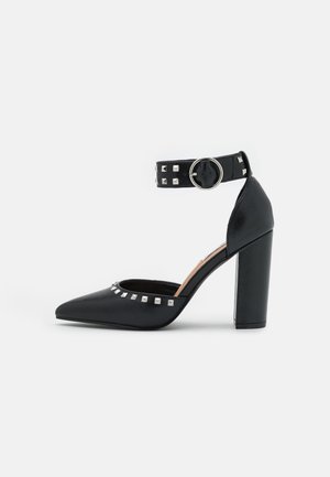 Chaussure noire à talon haut avec bout pointu, bride à la cheville ornée de clous argentés, et talon épais. Texture en cuir lisse et design minimaliste.