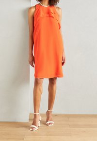 Robe orange sans manches avec un décolleté à volants, une coupe ample et une longueur au genou ; portée avec des talons blancs à brides. Texture lisse.