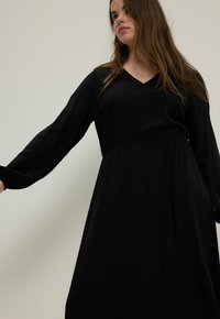 Vestido maxi negro con mangas largas y escote en V. Tejido suave, ceñido en la cintura, falda fluida, diseño minimalista. Sin patrones ni adornos.