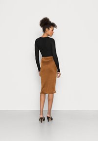 Blusa preta de manga longa combinada com uma saia de cetim bronze até ao joelho. A modelo está de pé em mules pretas de salto alto sobre um chão cinza claro.