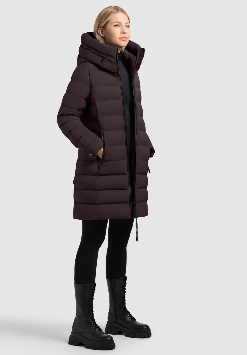 Puffer coat bordeaux con colletto alto, cuciture orizzontali, tasche con zip e forma leggermente svasata, indossato sopra leggings neri e stivali.
