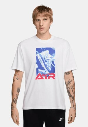 Giovane uomo con capelli rossi corti che indossa una maglietta bianca con grafica di sneaker blu e rossa e pantaloni neri Nike, in piedi davanti a uno sfondo semplice.