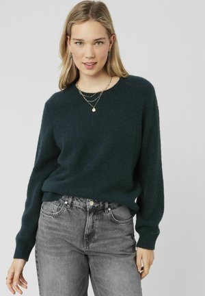 Pull en maille vert foncé, à manches longues et col rond. Porté sur un jean gris, il présente une coupe décontractée et une texture douce.