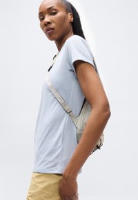 Γυναίκα με μακριά μαλλιά, που φοράει φωτεινό μπλε μπλουζάκι και μπεζ παντελόνι, με μπεζ τσάντα χιαστί, κοιτάζει με αυτοπεποίθηση πλάγια σε λευκό φόντο.