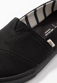 Svart kanvas slip-on sko med en texturerad yta, försedd med ett randigt tygfoder och en liten vit etikett på sidan.