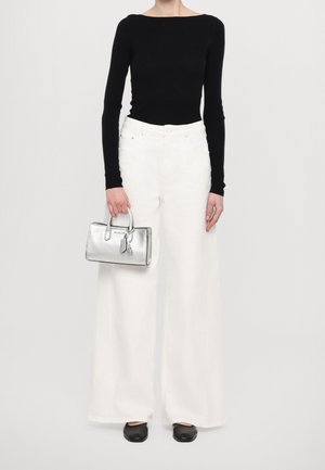 Person wearing black long-sleeve top and white wide-leg pants holding a silver handbag and wearing black shoes, standing against white background.