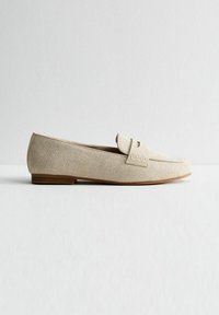 Mocasines beige texturizados con un acento de costura en la parte frontal y una suela plana marrón, que cuenta con una tira de cuero suave a lo largo del empeine.