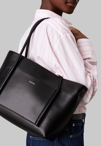 Borsa a tracolla nera in pelle con due manici lunghi e tasca frontale. Presenta un logo sottile sulla superficie. Indossata da una persona con una camicia a righe.