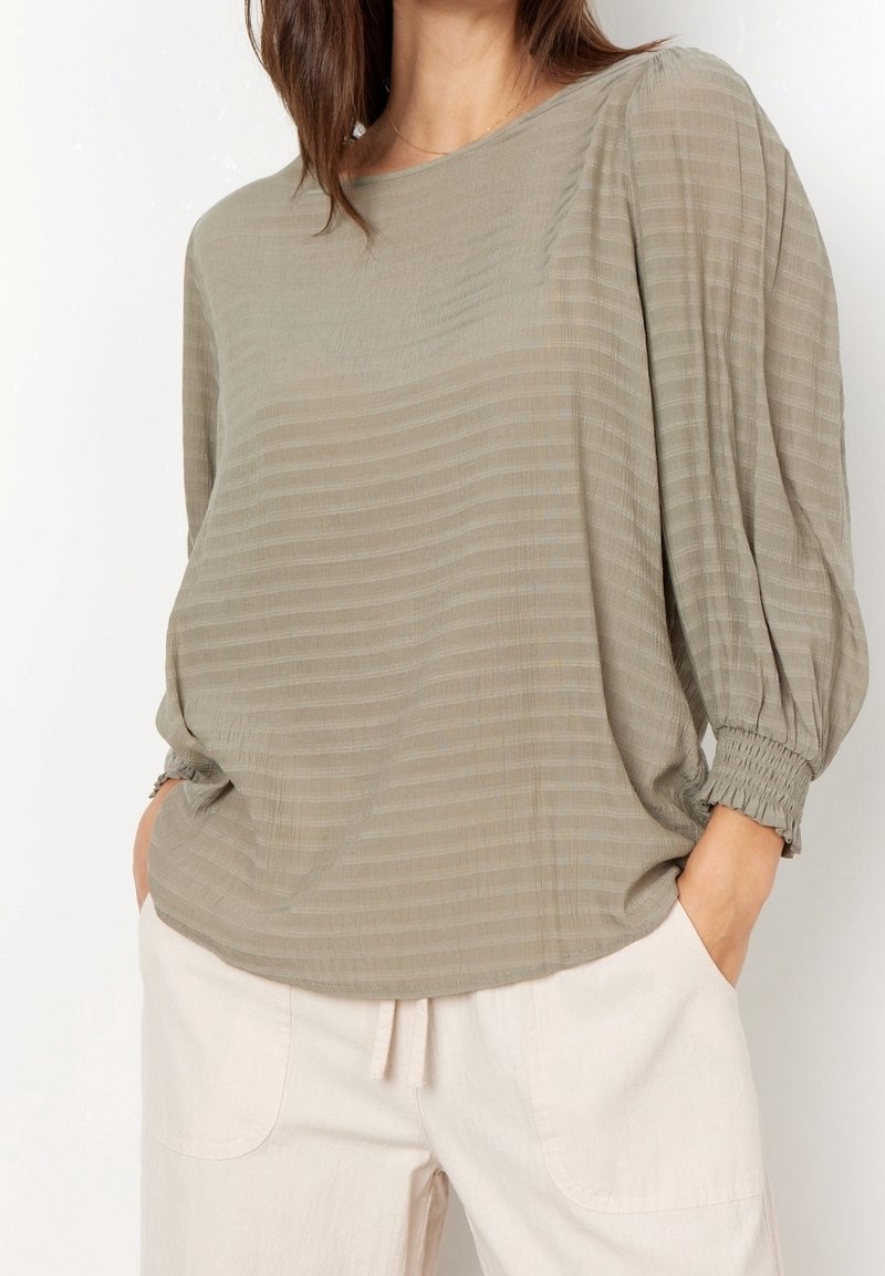 Blouse vert olive avec des rayures horizontales, coupe ample et poignets froncés. Tissu léger ; associée à un pantalon clair à coupe décontractée.