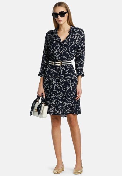 Rochie bleumarin cu model de text cursiv alb, mâneci lungi, închidere cu nasturi, talie strânsă cu un curea, asortată cu pantofi aurii și o poșetă albă.