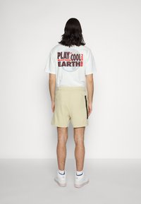 Vit t-shirt med grafisk text på baksidan, beige shorts med elastisk midja och sidoficka med dragkedja, i kombination med vita sneakers.