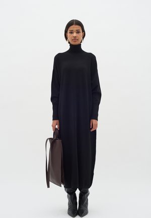 Black long-sleeve turtleneck dress in a loose fit, paired with knee-high black boots and a brown leather tote bag.