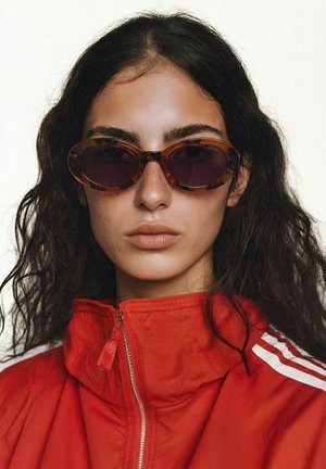 Jeune femme aux cheveux ondulés foncés portant des lunettes de soleil ovales à monture écaille de tortue et une veste zippée rouge avec des rayures blanches.