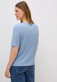 Een vrouw staat met haar rug naar ons gekeerd, gekleed in een lichtblauw shirt met korte mouwen en een spijkerbroek, met steil bruin haar, tegen een effen witte achtergrond.