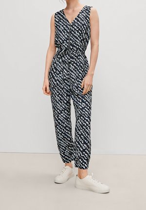 Femme portant une combinaison noire sans manches avec un motif abstrait blanc, une fermeture éclair à l'avant, une taille nouée et des baskets blanches sur fond uni.