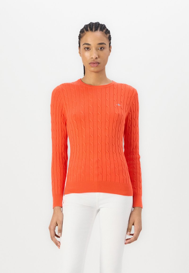 Pull en maille torsadée orange avec un col rond, des manches longues, des poignets et un ourlet côtelés. Petit logo sur la poitrine. Texture lisse.