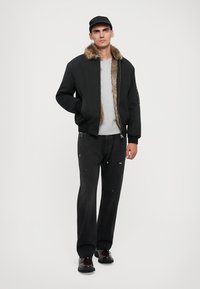Svart bomberjacka med faux pälskrage, grå t-shirt under, bärs med nött svarta jeans och svarta skor, beige keps på huvudet.