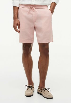 Mand iført lyserøde shorts med snøre og beige snørestøvler, stående med hænderne i lommerne mod en hvid baggrund.