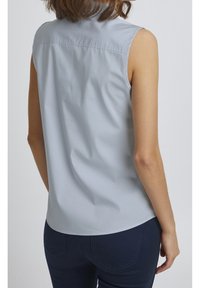 Jasnoniebieska bezrękawnikowa koszula z kołnierzykiem zapinanym na guziki, o gładkiej teksturze i detalach w tylnej części. Noszona z ciemnoniebieskimi dżinsami.