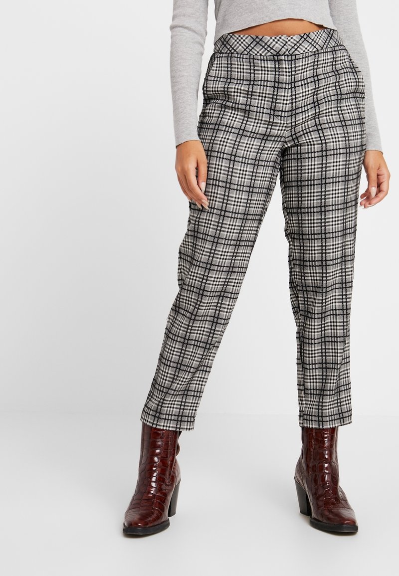 Femme portant un pantalon à carreaux gris, un haut court gris à manches longues et des bottines à talons marron, debout devant un fond clair.