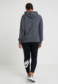 Mörkgrå huvtröja med en strukturerad yta, kombinerad med svarta leggings som har en stor vit Nike-logotyp på vänster sida.