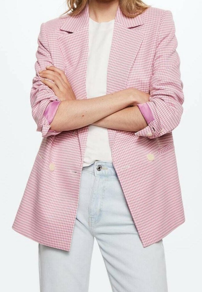 Personne portant un blazer pied-de-poule rose aux manches retroussées, une chemise blanche et un jean bleu clair, les bras croisés au niveau de la poitrine.