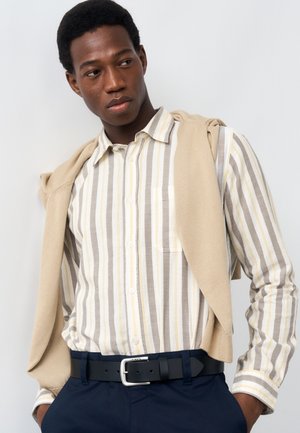 Άνδρας που φοράει πουκάμισο με ρίγες σε μπεζ και λευκό, με μπεζ πουλόβερ περασμένο στους ώμους, σκούρο μπλε παντελόνι και μαύρη ζώνη, κοιτάζει αριστερά.