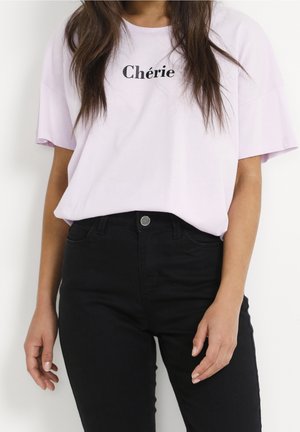 Kobieta ubrana w jasnofioletowy T-shirt z napisem „Chérie” oraz wysokowalne czarne dżinsy, stojąca na tle jednolitego białego tła.