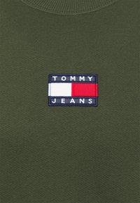 Tmavě zelená pletenina s obdélníkovou nášivkou loga Tommy Jeans v námořnické modři, bílé a červené, umístěnou centrálně pod kulatým výstřihem.