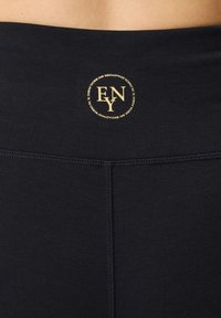 Leggings neri con una texture liscia, caratterizzati da un logo circolare dorato con la scritta "FNY" sulla vita. Cuciture piatte e un design aderente.