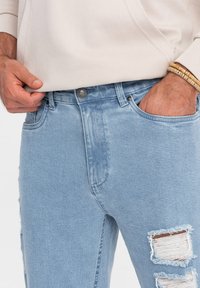 Jasnoniebieskie dżinsy z klasycznym pięciopocketowym wzorem, zapinane na guzik z przodu, ozdobione przetarciami i postrzępionymi brzegami.