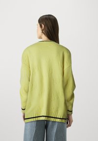 Sweater oversized amarelo claro com padrão de texto em relevo, bainha listrada em preto e gola canelada, usado sobre calças de denim azuis.