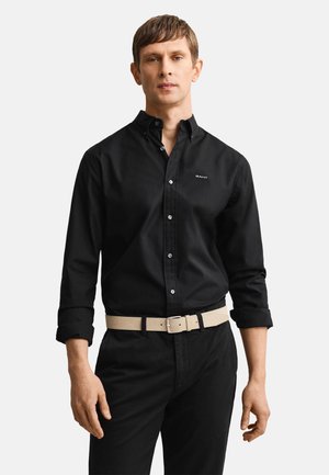 Mężczyzna w czarnej koszuli na guziki z podwiniętymi rękawami, czarnych spodniach i beżowym pasku, stoi na tle jednolitego jasnego tła.