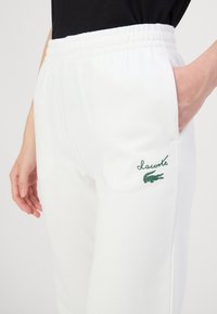 Baltos sportinės kelnės su elastingu juosmeniu, šoninėmis kišenėmis ir žaliu Lacoste logotipu kairėje šlaunyje. Minkšta medžiaga ir laisvas siluetas.