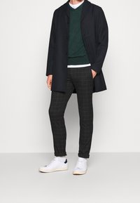 Homme portant un manteau noir boutonné, un pull vert sur une chemise blanche, un pantalon à carreaux foncé, des chaussettes blanches et des baskets blanches, debout devant un fond uni.