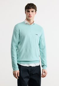 Jeune homme portant un pull à manches longues vert menthe avec le logo "BOSS", une chemise blanche à col en dessous, et un pantalon noir, debout devant un fond blanc.