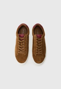 Zapatillas de ante marrón con una suela de goma blanca, que presentan cordones planos, acentos en burdeos en el cuello y un logo grabado en la lengüeta.