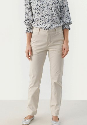 Beige Chinos mit glatter Textur, mittelhohem Bund, seitlichen Taschen und geradem Bein, kombiniert mit einer blumenbedruckten Bluse.