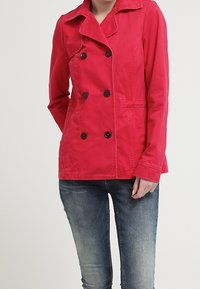 Abrigo corto rojo con diseño de doble botonadura, botones negros y cuello texturizado, combinado con vaqueros azules. La tela parece suave y ligera.