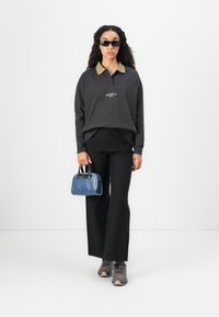 Kvinne med langt, krøllete hår som har på seg svarte solbriller, mørk langermet polo, svarte bukser, grå joggesko, og holder en liten blå veske.